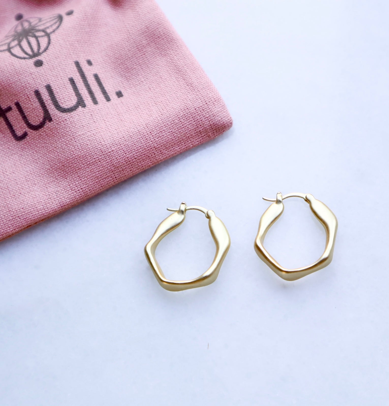 Gold hoop earrings on a white surface with a pink pouch labeled 'tuuli' in the background.
